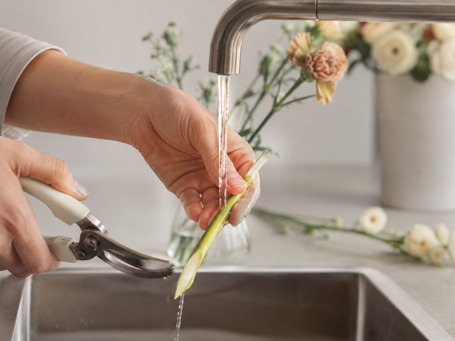 Florist recutting a flower stem at a sharp angle under running water, the single most important step to making a bouquet last longer