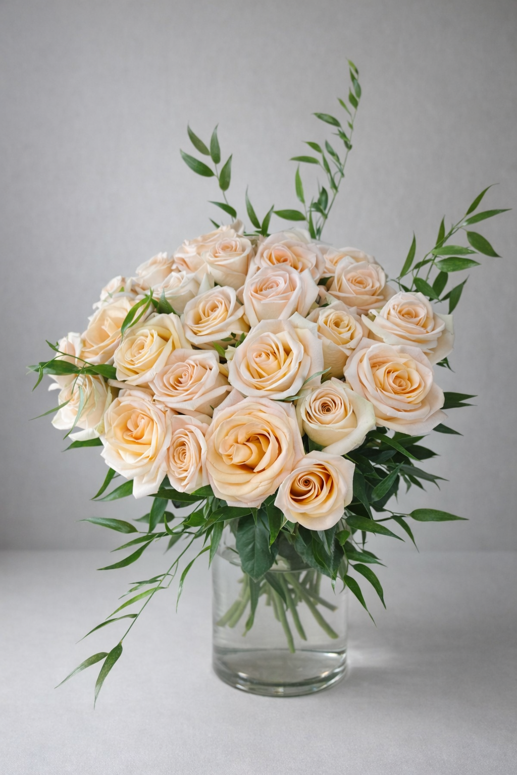 Peach rose bouquet in a clear glass vase with fresh greenery, photographed front-facing in a soft editorial studio setting.