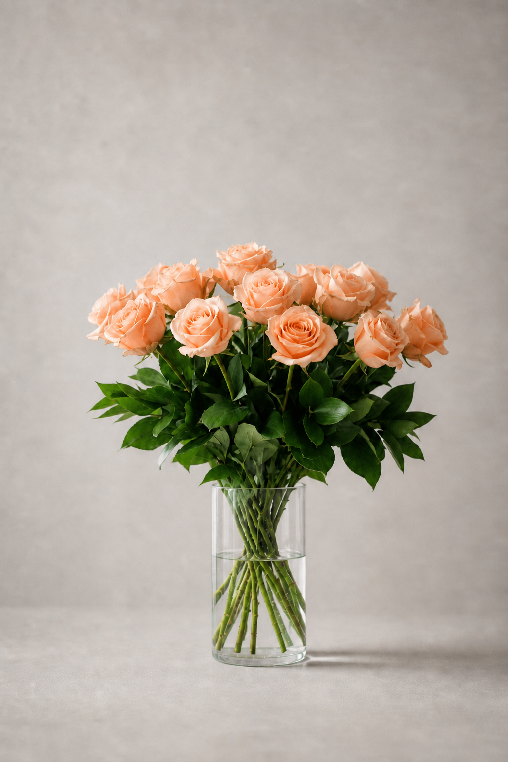 Peach rose bouquet in a clear glass vase with fresh greenery, photographed front-facing in a soft editorial studio setting.