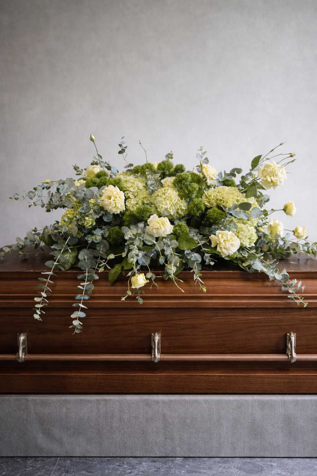 Elegant sympathy casket spray with cream lisianthus, green hydrangeas, green trick dianthus and eucalyptus displayed on a walnut coffin in a refined editorial setting.