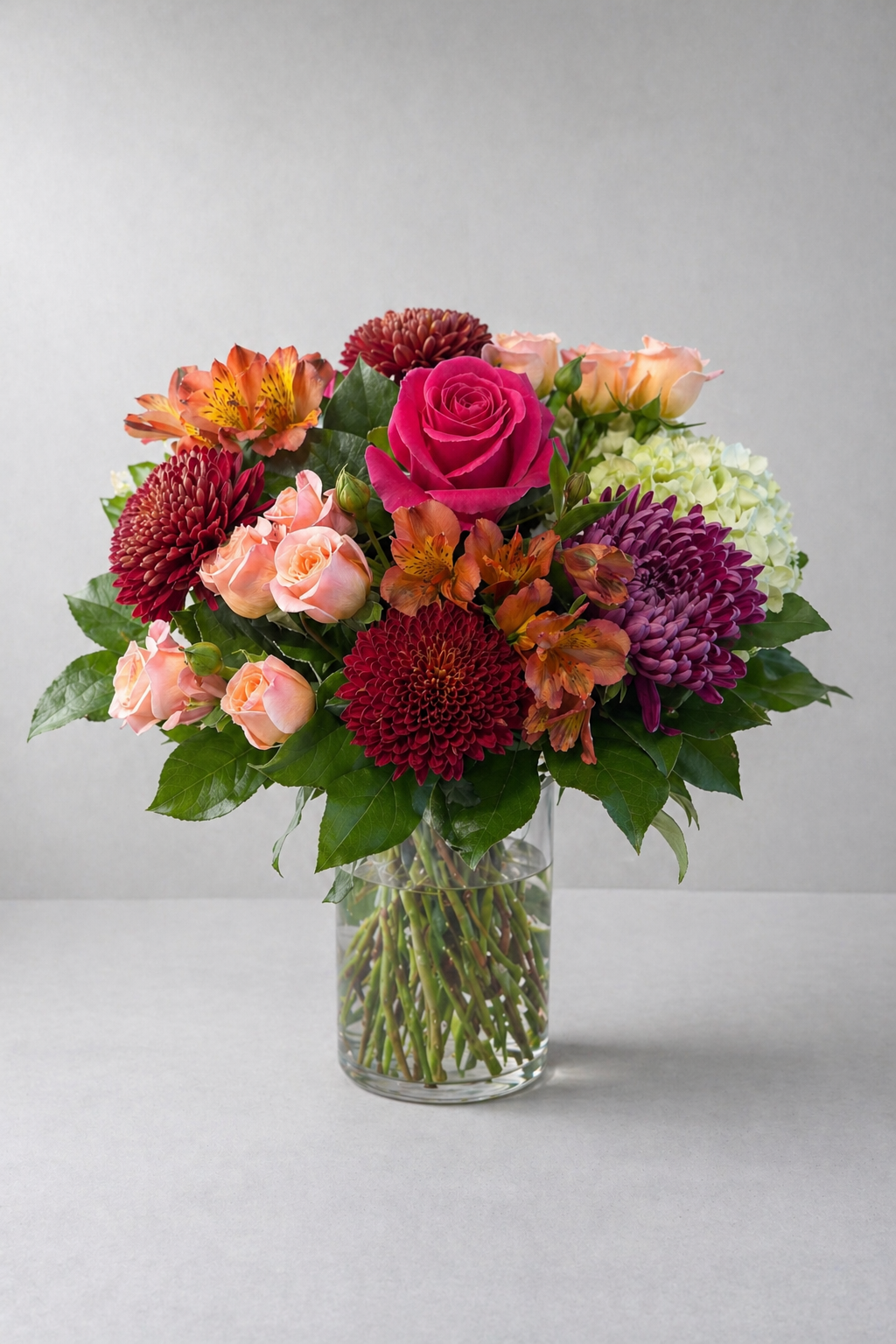 Luxury autumn bouquet in a clear glass vase with a vivid pink rose, peach spray roses, burgundy pompon chrysanthemums, mauve chrysanthemum, orange alstroemeria and pale green hydrangea, photographed front-facing in a refined editorial style by a Montreal florist.