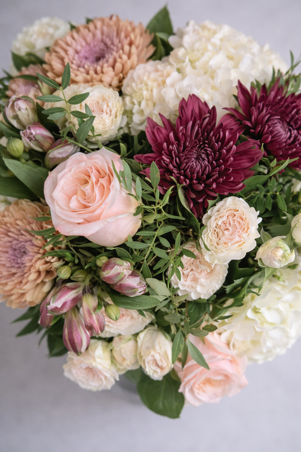 Top view of a refined floral arrangement with blush roses, burgundy chrysanthemums, cream spray roses, white hydrangeas and fresh greenery