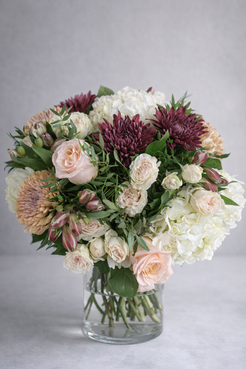 hree-quarter view of a luxury flower arrangement with blush roses, burgundy chrysanthemums, white hydrangeas and cream spray roses in a clear vase