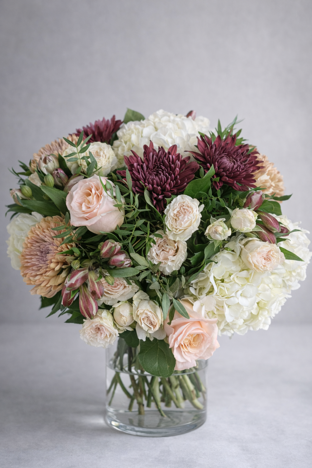 hree-quarter view of a luxury flower arrangement with blush roses, burgundy chrysanthemums, white hydrangeas and cream spray roses in a clear vase