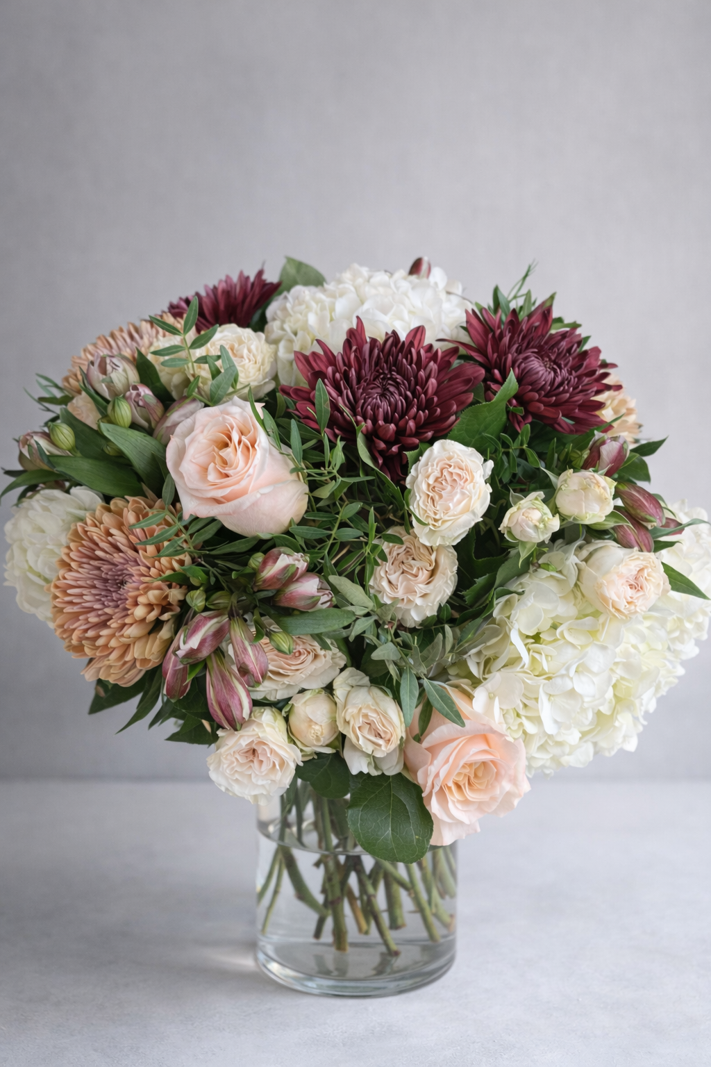 Luxury bouquet with blush roses, burgundy chrysanthemums, white hydrangeas and cream spray roses in a clear glass vase, front view