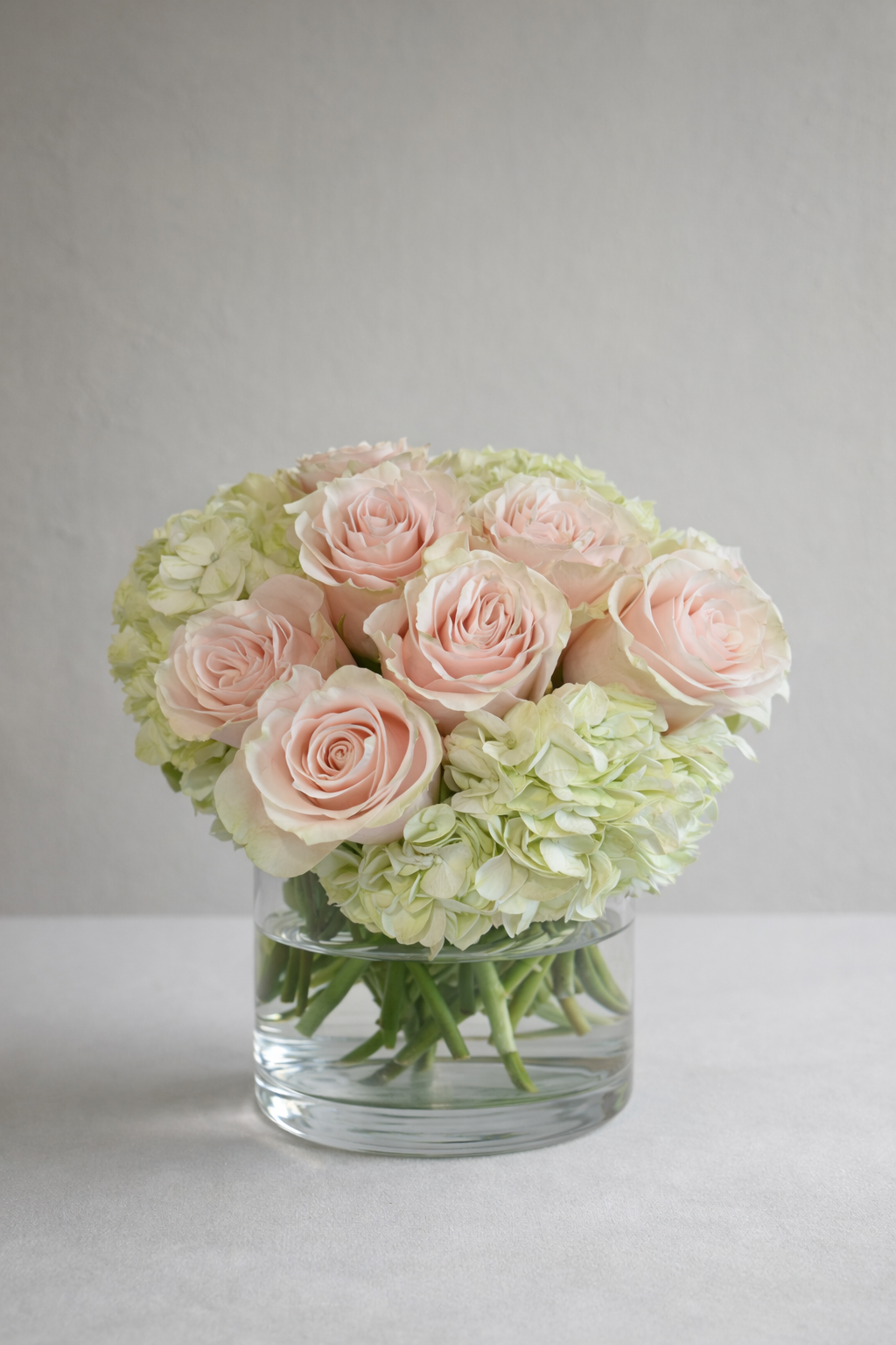 Pale blush roses and soft green hydrangeas arranged in a rounded clear glass vase, photographed straight on with balanced symmetry for an elegant floral arrangement Montreal by a luxury florist Montreal.