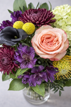 Botanical close-up top view of a refined bouquet showing peach rose petals, dark calla lily, purple alstroemeria, burgundy chrysanthemums and textured foliage.