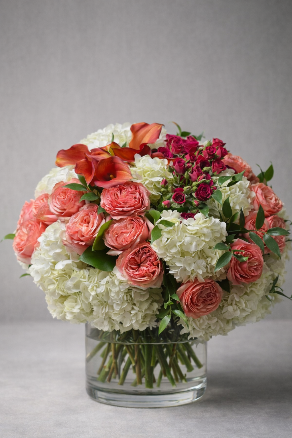 Luxury floral arrangement with coral garden roses, white hydrangeas, magenta spray roses, and orange calla lilies in a clear glass cylinder vase, photographed front-facing in an editorial style by a Montreal florist.