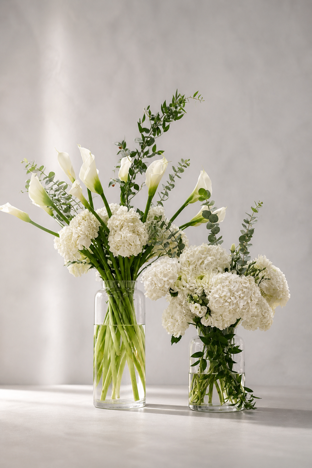 Editorial lifestyle image of funeral urn flowers in a warm contemporary interior, with white calla lilies, hydrangeas and foliage in clear glass vases beside a modern urn on a refined console, by a luxury florist Montreal.