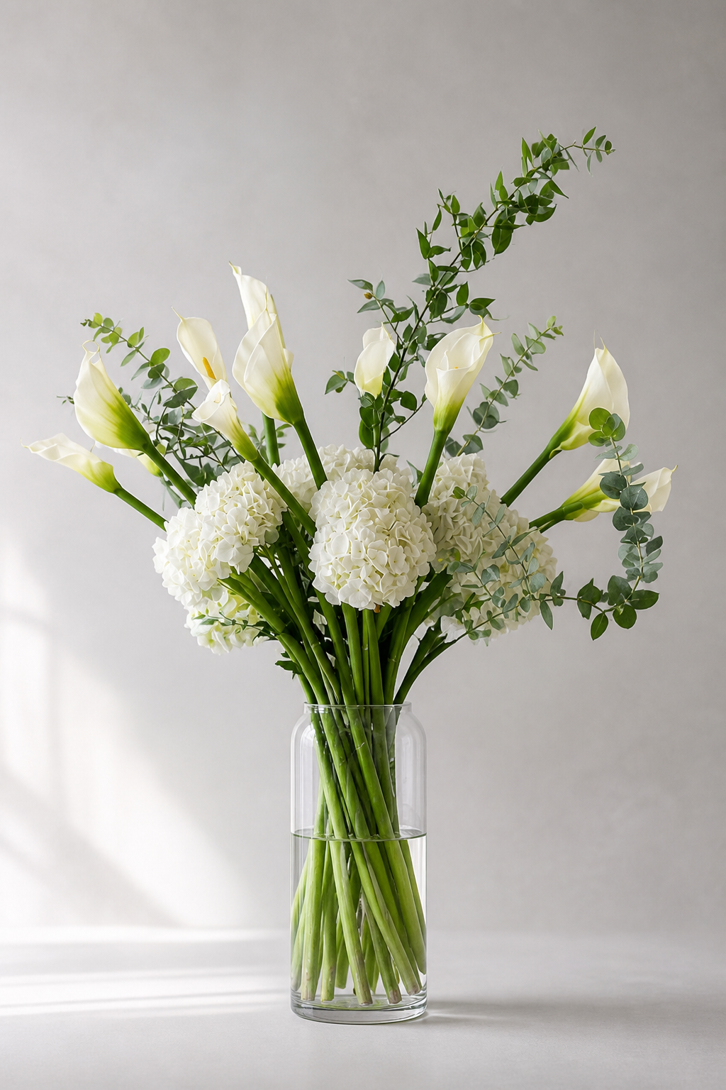 One vase of set of two urn flower tribute with white calla lilies, hydrangeas, eucalyptus and Italian ruscus in matching clear vases framing a modern urn, designed by a Montreal florist.