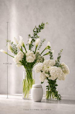 Size reference image of a premium funeral urn flower arrangement with white calla lilies, hydrangeas, eucalyptus and Italian ruscus in clear cylindrical vases beside a modern urn, styled for a Montreal florist collection.