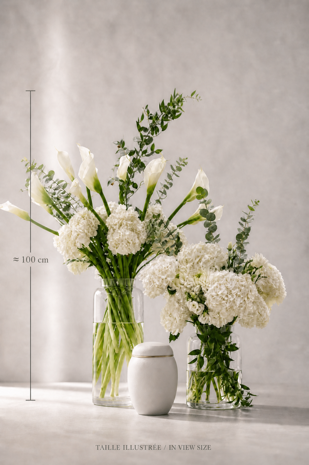 Size reference image of a premium funeral urn flower arrangement with white calla lilies, hydrangeas, eucalyptus and Italian ruscus in clear cylindrical vases beside a modern urn, styled for a Montreal florist collection.