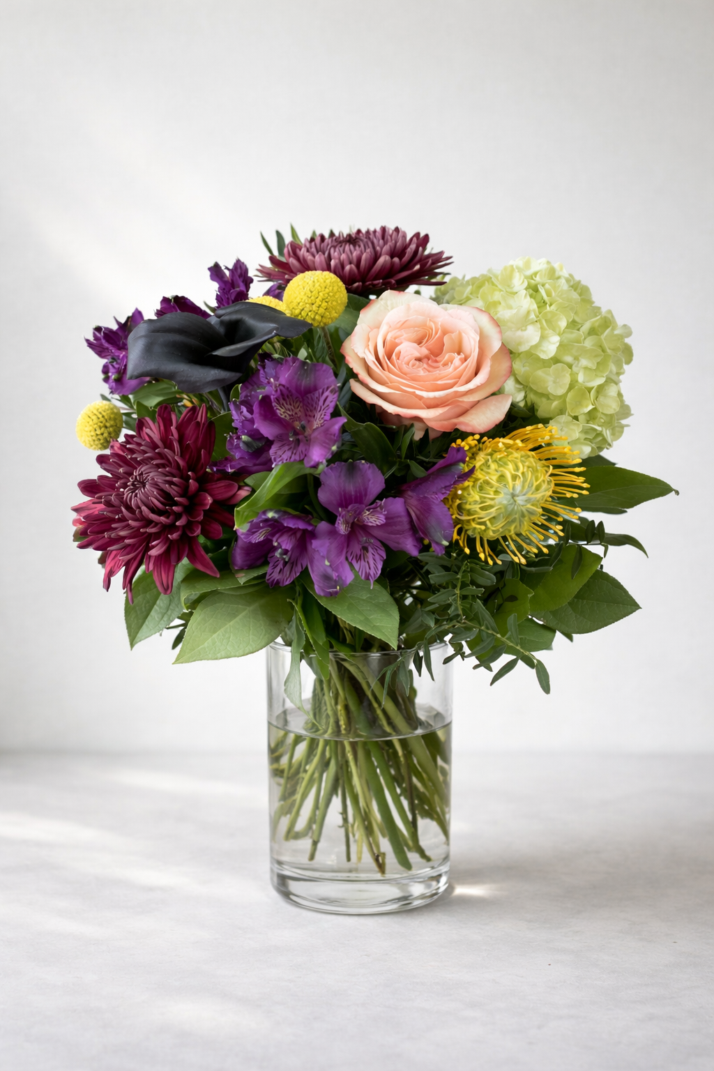 Luxury editorial bouquet with peach rose, dark calla lily, burgundy chrysanthemums, purple alstroemeria and green hydrangea in a clear glass vase.