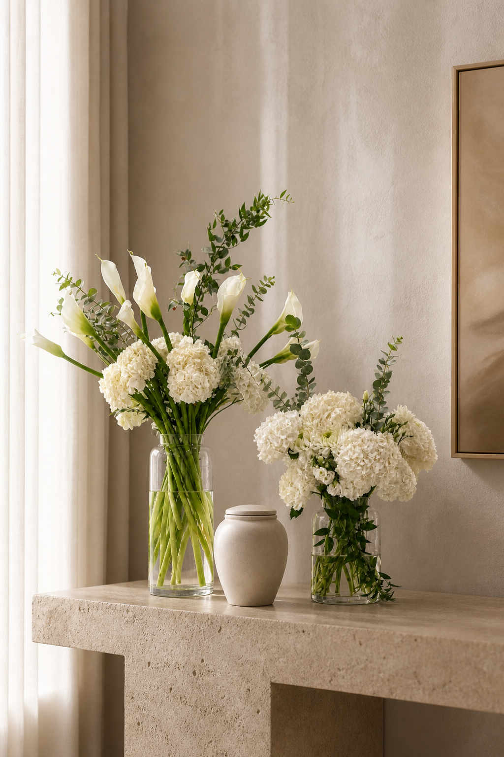 Modern asymmetrical funeral urn flower design with a taller white calla lily arrangement and a lower hydrangea arrangement beside a contemporary urn, photographed in a soft luxury interior for flower delivery Montreal.