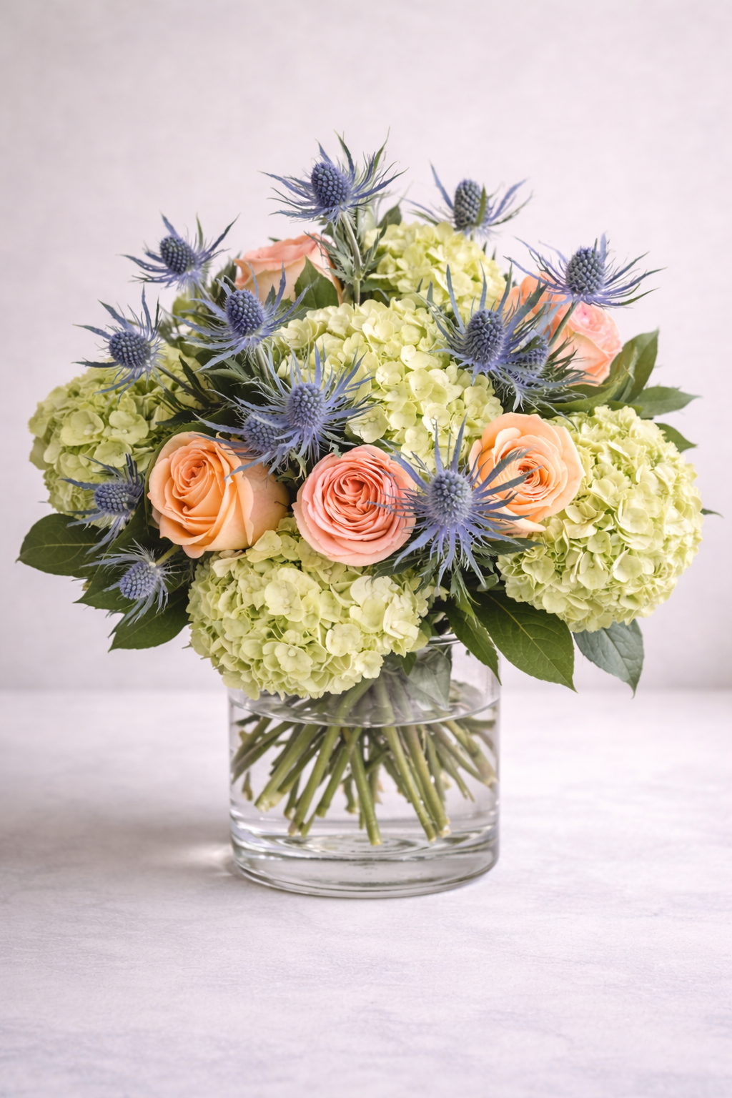 Luxury bouquet with peach roses green hydrangea and blue eryngium in glass vase angled view Montreal flower delivery
