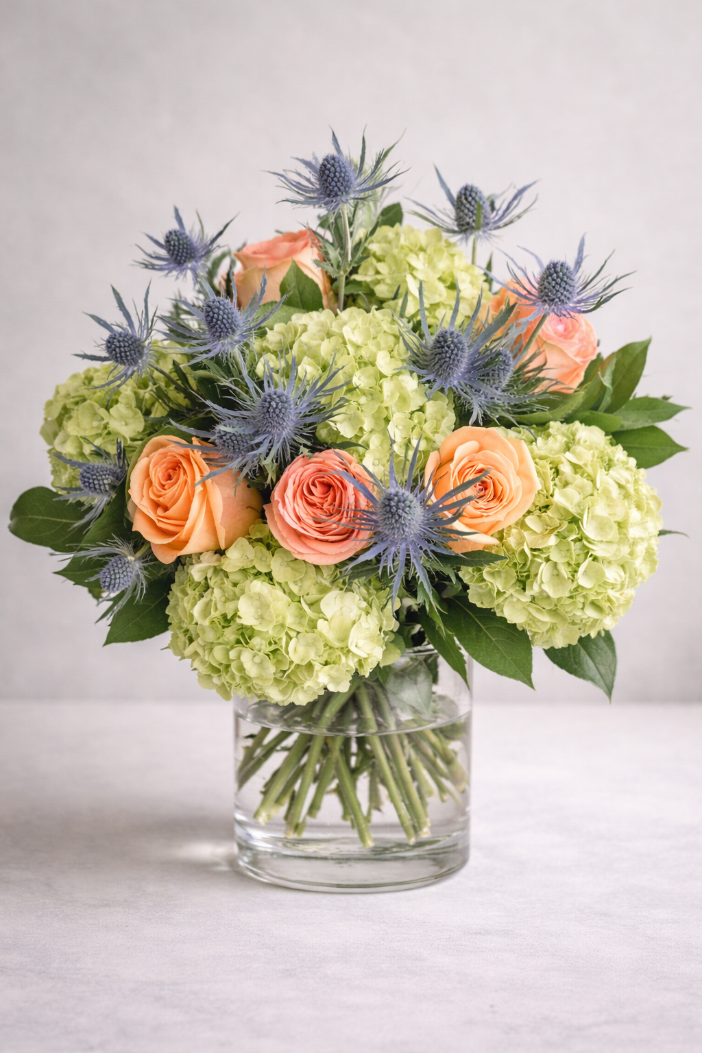 Peach roses with green hydrangea and blue eryngium bouquet in clear glass vase by Montreal luxury florist