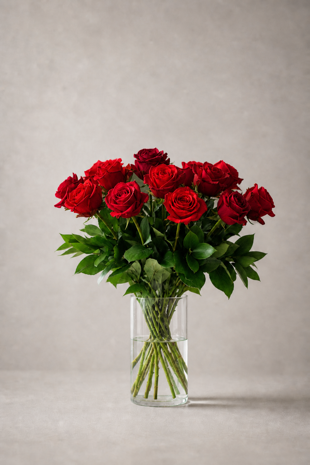 Red Ecuador Freedom roses arranged in a rounded luxury bouquet with lush green foliage in a clear cylindrical glass vase, photographed front-facing against a soft pearl-grey editorial backdrop by a Montreal florist.