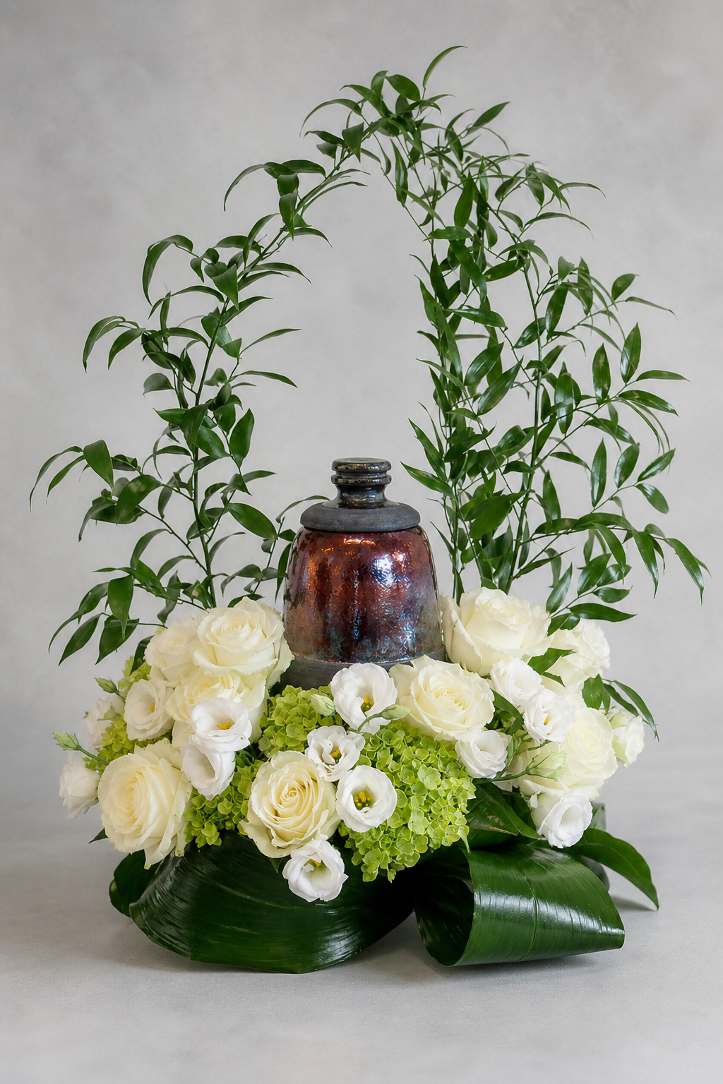Luxury urn floral tribute with white roses, white lisianthus, green hydrangea and sculptural glossy foliage, photographed front-facing in a refined editorial style by a Montreal florist.