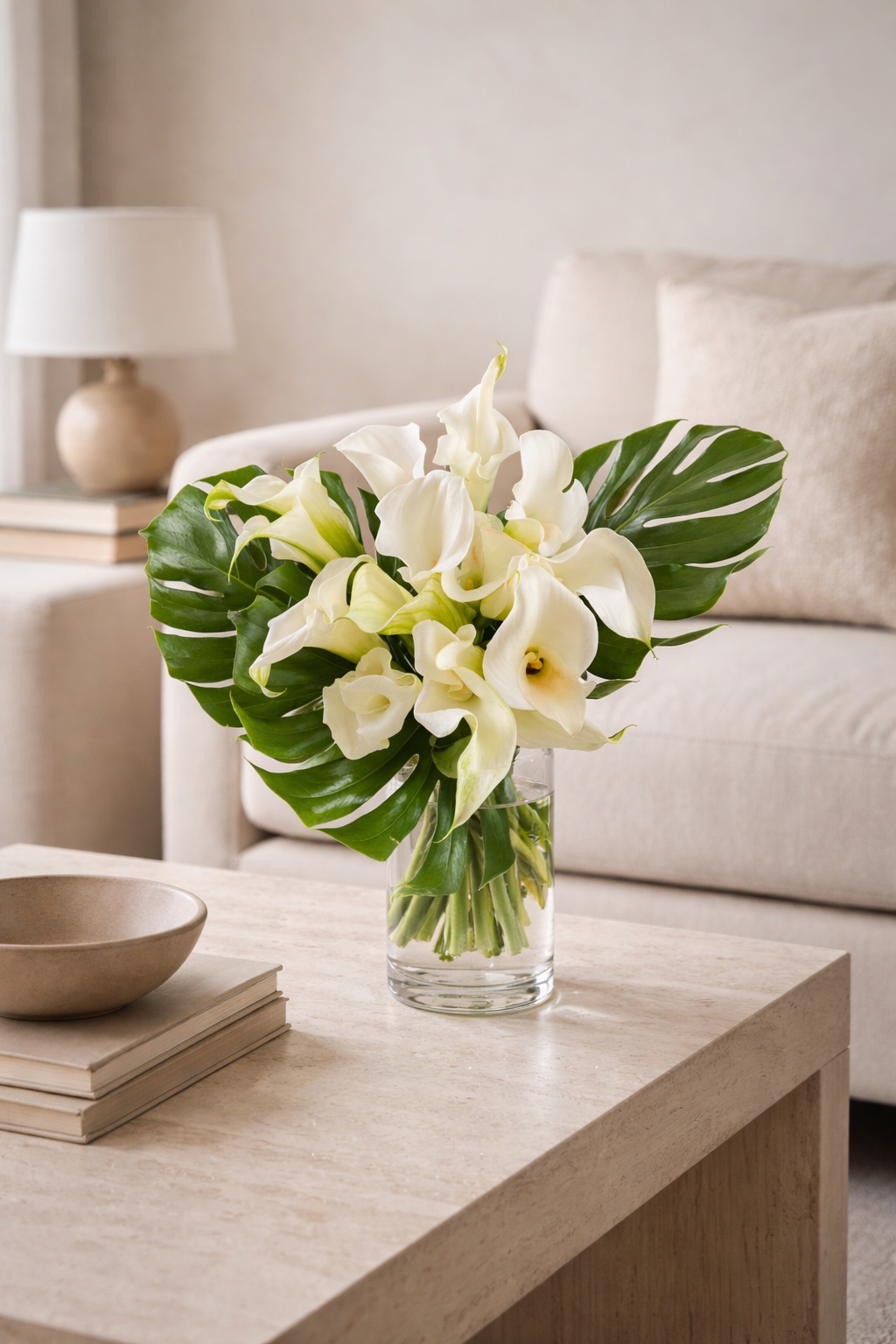 White calla lily bouquet styled in a contemporary neutral interior, arranged in a clear glass vase with monstera leaves, Montreal florist