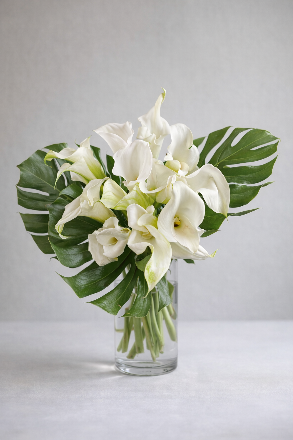 White calla lily bouquet with monstera leaves in a clear glass vase, photographed in a refined editorial style by a Montreal florist

