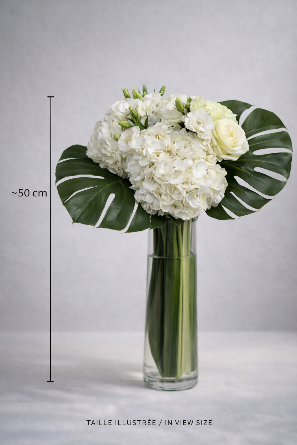 White floral arrangement with hydrangeas, roses, lisianthus and monstera leaves in a tall clear glass vase, shown at approximately 50 cm for size reference, an elegant premium bouquet Montreal design.