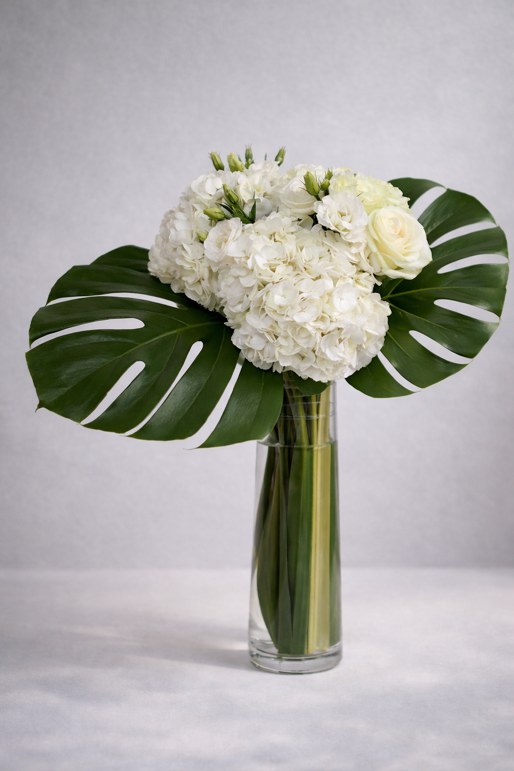 White hydrangea, rose and lisianthus bouquet in a clear cylindrical vase with bold monstera leaves, photographed front-facing in a refined editorial style by a Montreal florist.